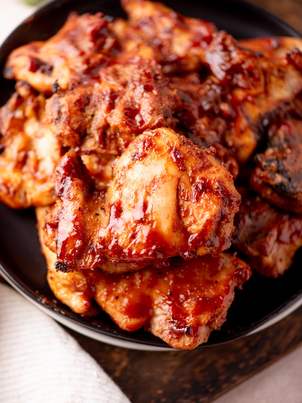 Close-up of several pieces of grilled chicken coated in a glossy, reddish barbecue sauce, stacked on a dark plate with a white cloth partially visible underneath.