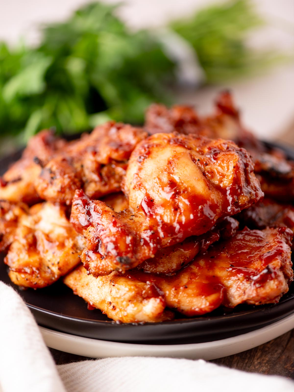 A close-up of a plate piled with glazed, grilled chicken pieces, garnished with herbs in the background. The chicken appears juicy and covered in a sticky, reddish sauce.
