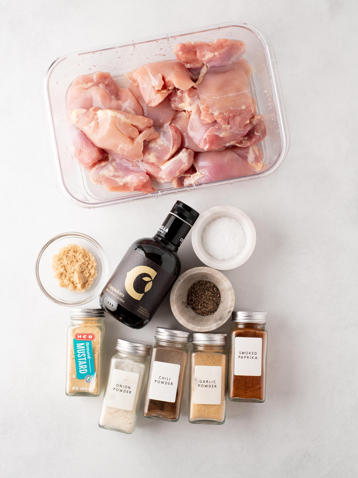 Raw chicken thighs in a glass container, surrounded by bowls and jars of ingredients including olive oil, salt, pepper, brown sugar, and various spices like mustard, onion powder, chili powder, garlic powder, and smoked paprika.