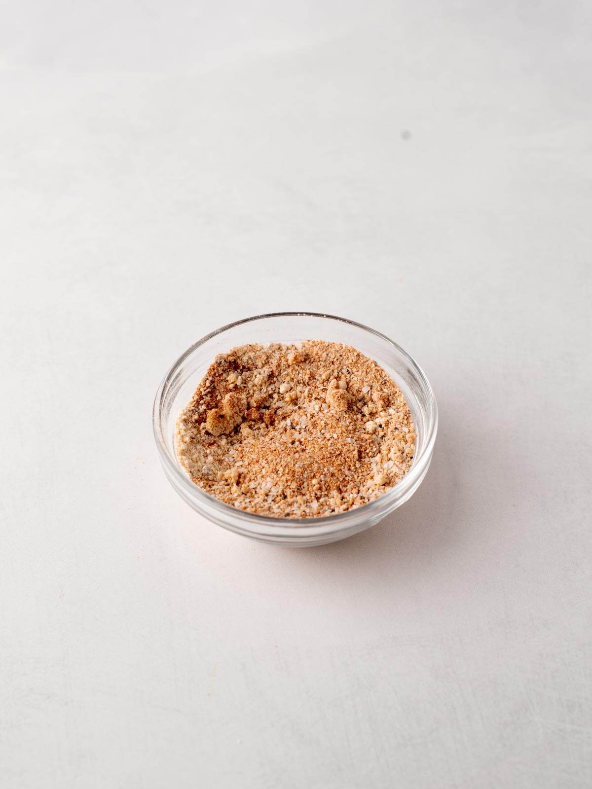 A small glass bowl filled with a light brown, finely ground seasoning mix sits on a plain, light-colored surface.