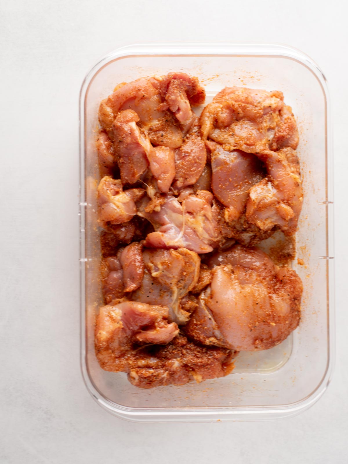 Raw chicken pieces coated in a reddish-brown spice marinade are arranged in a clear plastic container on a light-colored surface.
