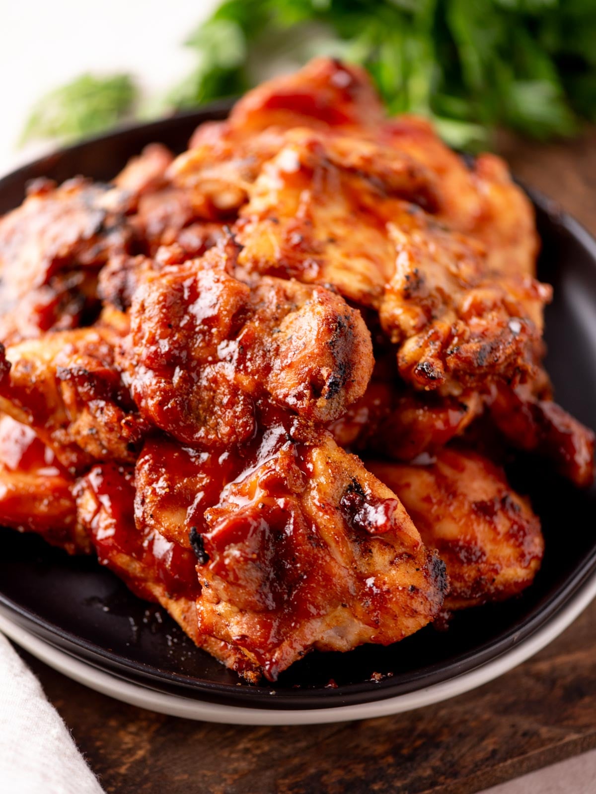 A close-up of a plate piled with grilled, saucy barbecue chicken thighs, glistening with a red, smoky glaze. Fresh parsley is blurred in the background.