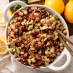 A white bowl filled with a colorful bean salad featuring chickpeas, kidney beans, artichokes, sun-dried tomatoes, diced celery, and herbs. Sliced lemons are next to the bowl on a wooden surface. Gold utensils rest inside the bowl.