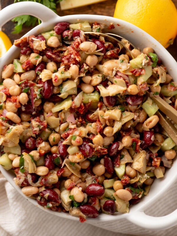 A white bowl filled with a colorful bean salad featuring chickpeas, kidney beans, artichokes, sun-dried tomatoes, diced celery, and herbs. Sliced lemons are next to the bowl on a wooden surface. Gold utensils rest inside the bowl.