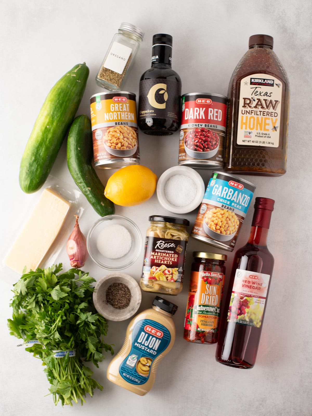 A flat lay of various ingredients, including canned beans, cucumber, lemon, honey, red wine vinegar, Dijon mustard, herbs, sun-dried tomatoes, cheese, black pepper, salt, olives, and a small bottle of balsamic vinegar.
