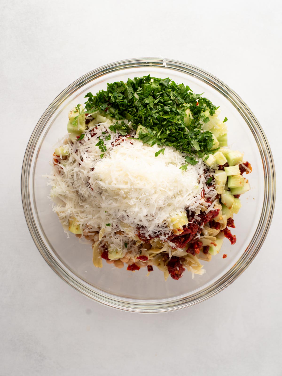 A glass bowl filled with chopped parsley, grated cheese, diced celery, sun-dried tomatoes, and artichoke hearts on a white background.