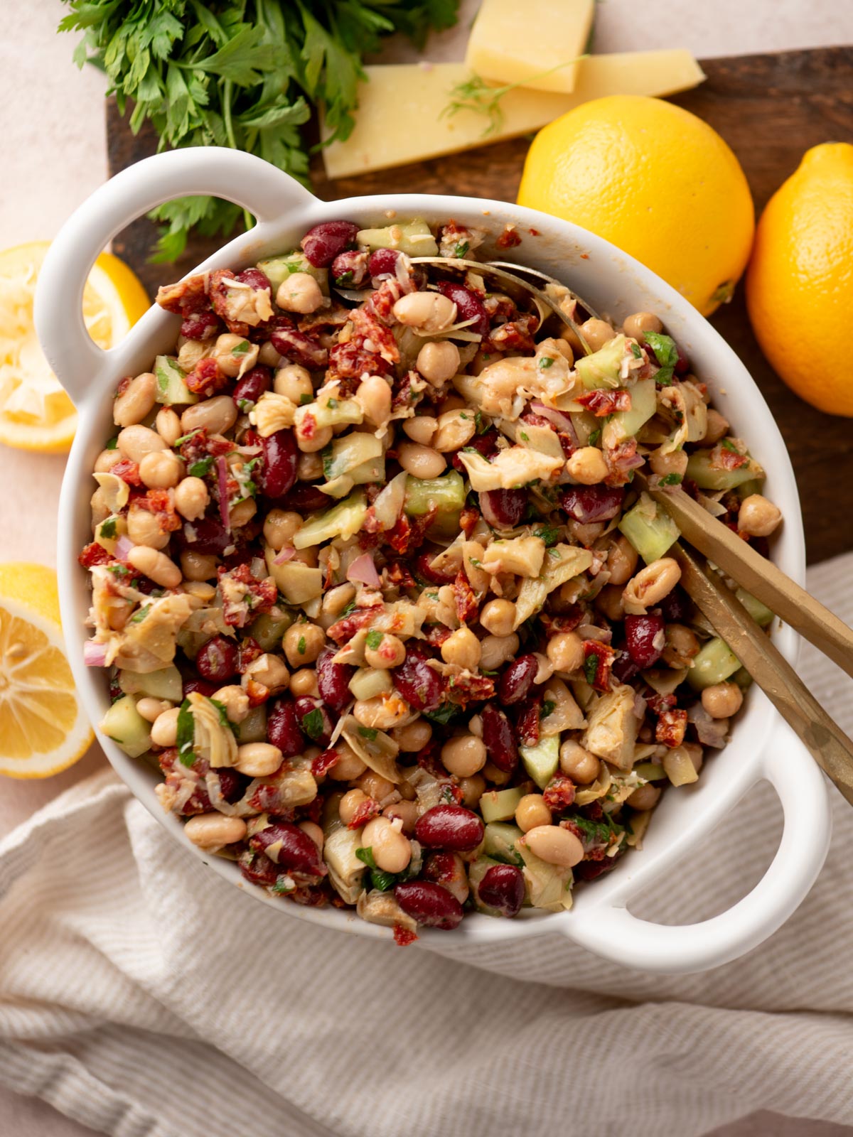 A white bowl filled with a colorful bean salad featuring kidney beans, chickpeas, white beans, chopped vegetables, and herbs. Lemons, cheese slices, and fresh parsley are on a wooden board in the background.