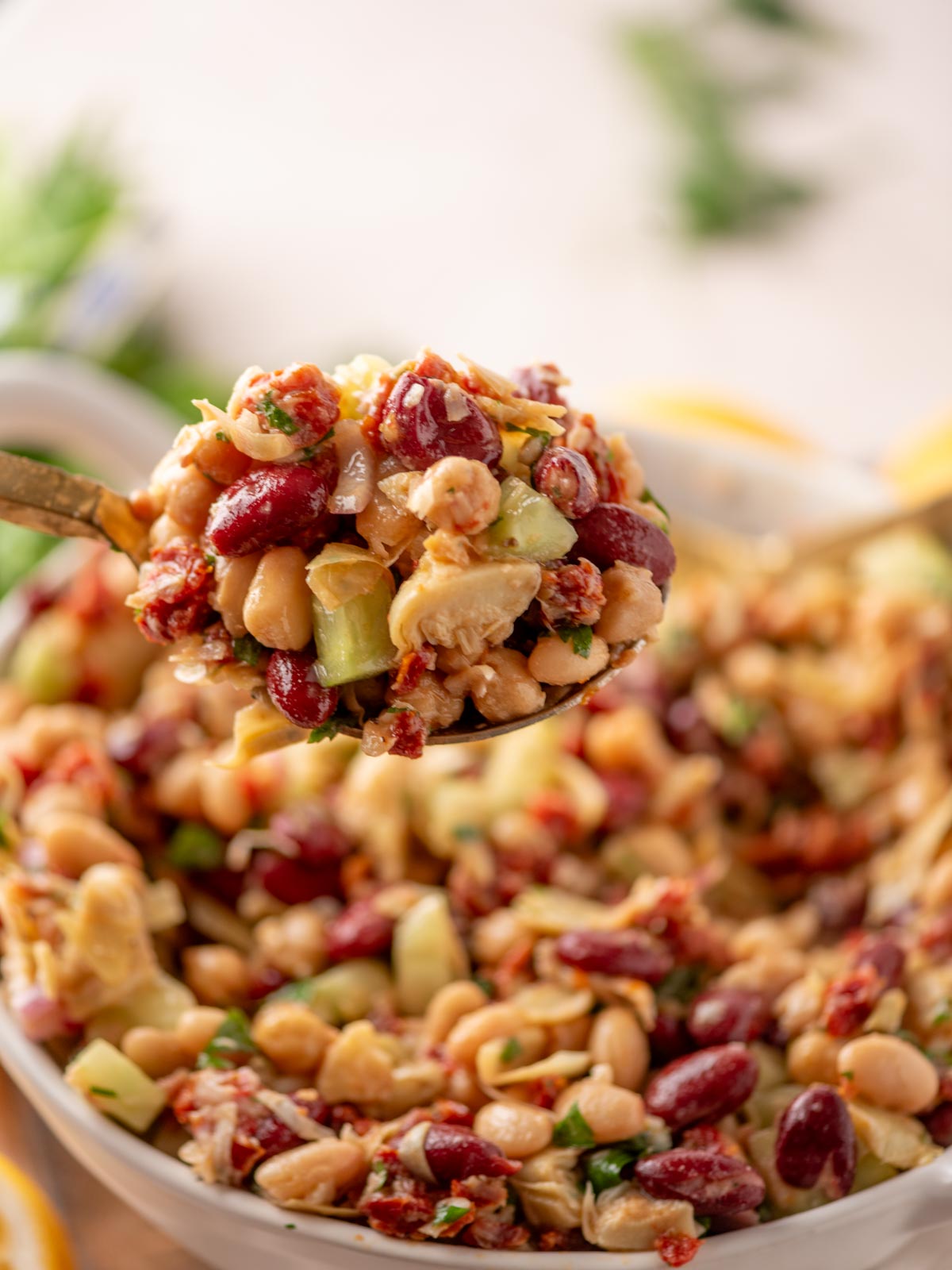 A spoonful of mixed bean salad with kidney beans, chickpeas, white beans, chopped cucumber, parsley, and sun-dried tomatoes, held above a bowl filled with the same salad.