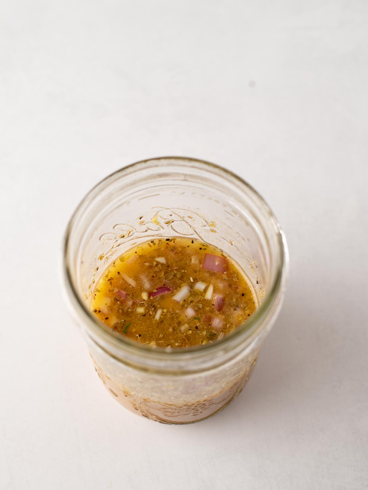 A glass jar filled with a chunky vinaigrette-style dressing containing visible bits of red onion and herbs, set against a plain white background.
