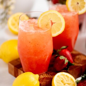 Two glasses of strawberry lemonade with lemon slices on the rim, surrounded by fresh strawberries and lemons on a wooden board. The drink is a bright pinkish-red color and appears cold and refreshing.