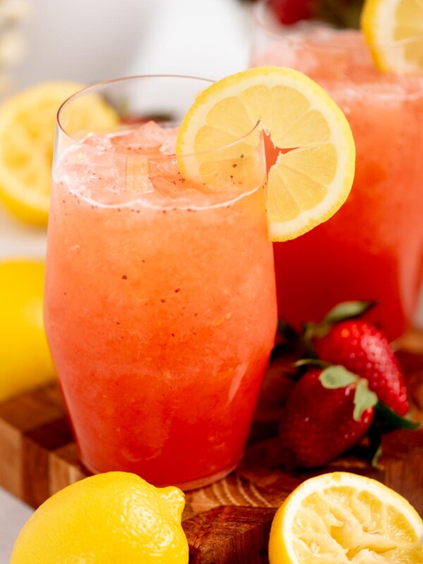 Two glasses of strawberry lemonade with lemon slices on the rim, surrounded by fresh strawberries and lemons on a wooden board. The drink is a bright pinkish-red color and appears cold and refreshing.