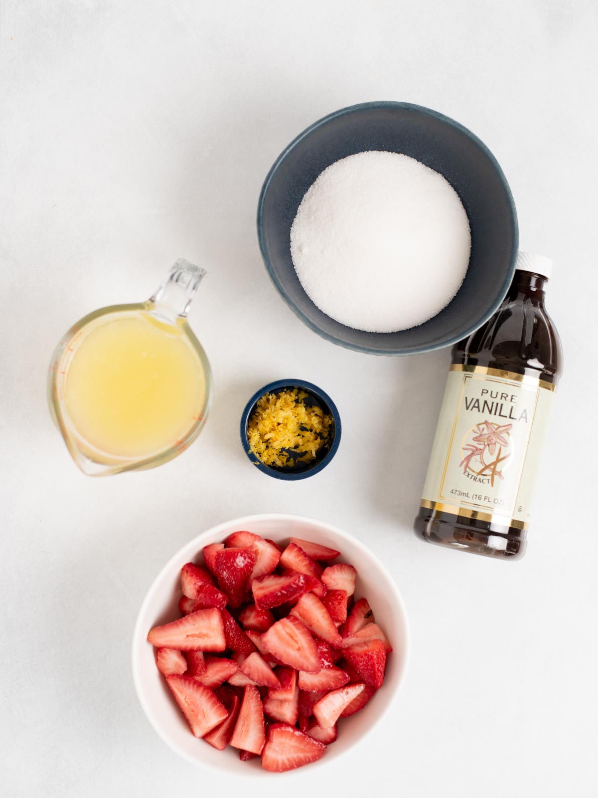 A bowl of sliced strawberries, lemon zest, lemon juice, sugar, and pure vanilla are arranged on a white surface—perfect for making fresh strawberry lemonade at home.