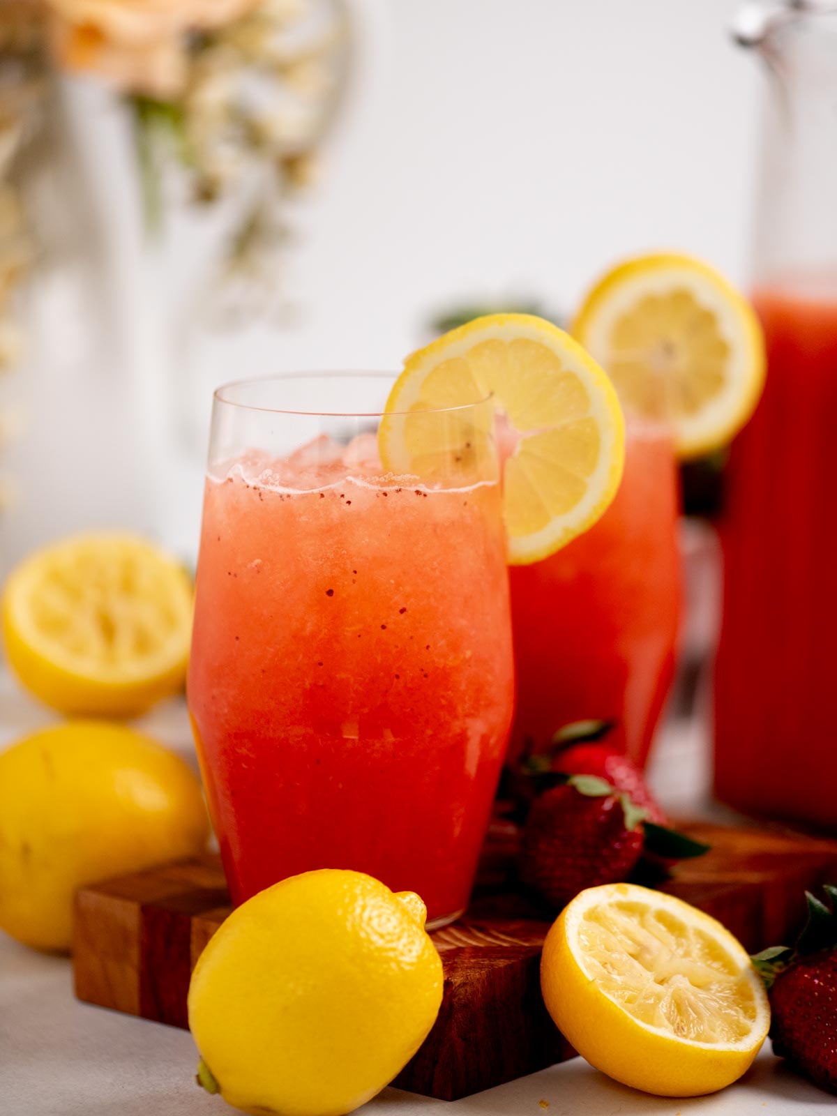 A tall glass of strawberry lemonade garnished with a lemon slice, surrounded by fresh lemons and strawberries on a wooden board, with another drink and blurred flowers in the background.