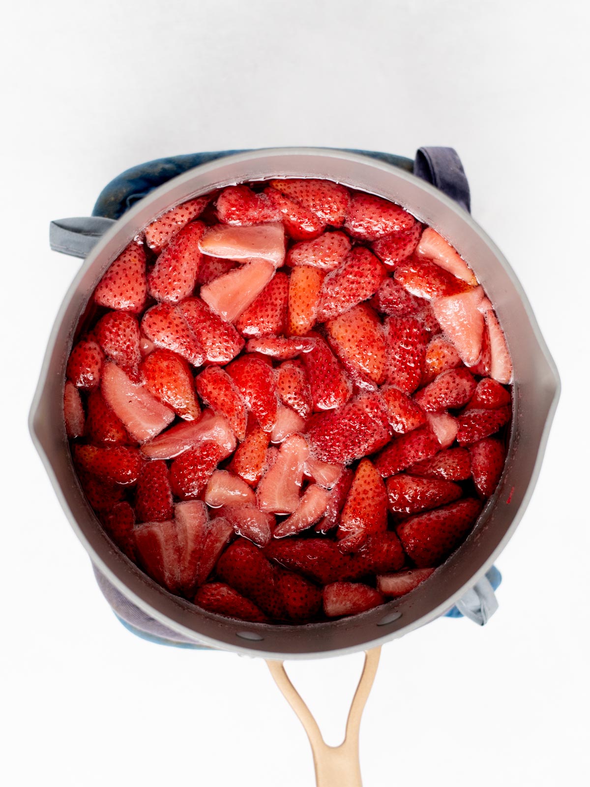 A pot filled with chopped strawberries sits on a blue oven mitt, viewed from above. The strawberries are bright red and appear to be cooking.