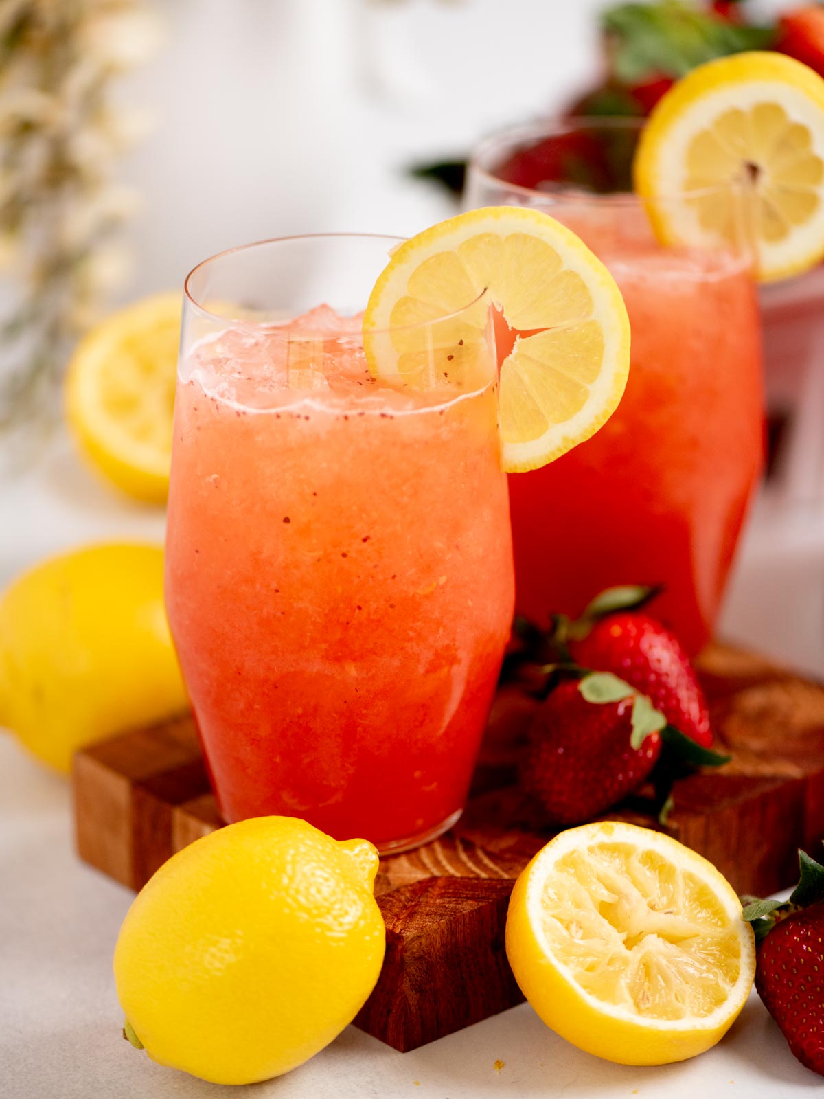 Two glasses of strawberry lemonade with lemon slices on the rims, surrounded by fresh strawberries and lemons, including a halved lemon, on a wooden board. The bright drinks look refreshing and vibrant.