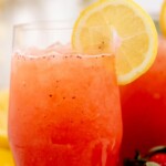 A glass of strawberry lemonade garnished with a lemon slice, surrounded by fresh lemons and strawberries, with a sign above reading strawberry lemonade and sweetlysplendid.com.