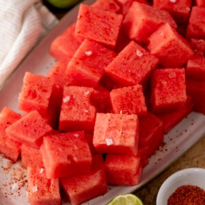 A close-up of a plate filled with cubed watermelon sprinkled with salt and chili powder, with a lime wedge and a bowl of chili powder nearby.