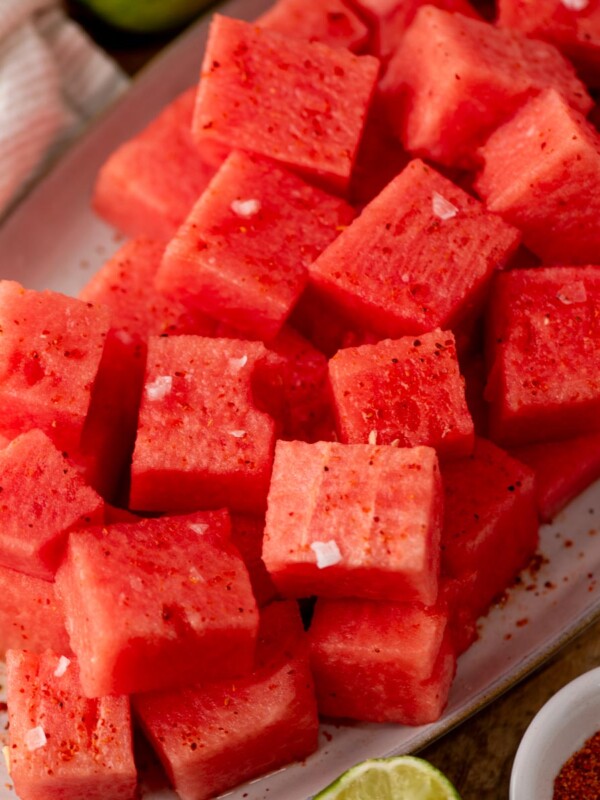 A close-up of a plate filled with cubed watermelon sprinkled with salt and chili powder, with a lime wedge and a bowl of chili powder nearby.