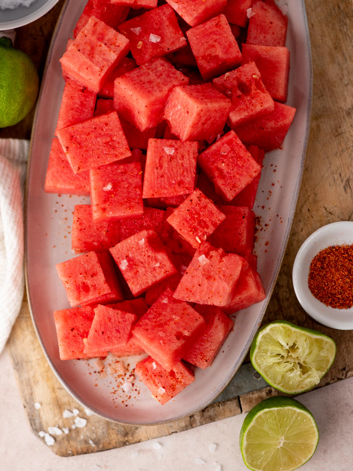 Cubed watermelon sprinkled with chili powder and coarse salt on a platter, surrounded by halved limes, a bowl of chili powder, and a wooden surface.