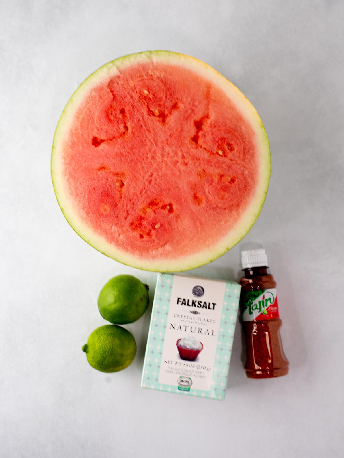 A halved watermelon, two limes, a box of Falksalt natural salt, and a bottle of Tajin seasoning are arranged on a white surface.