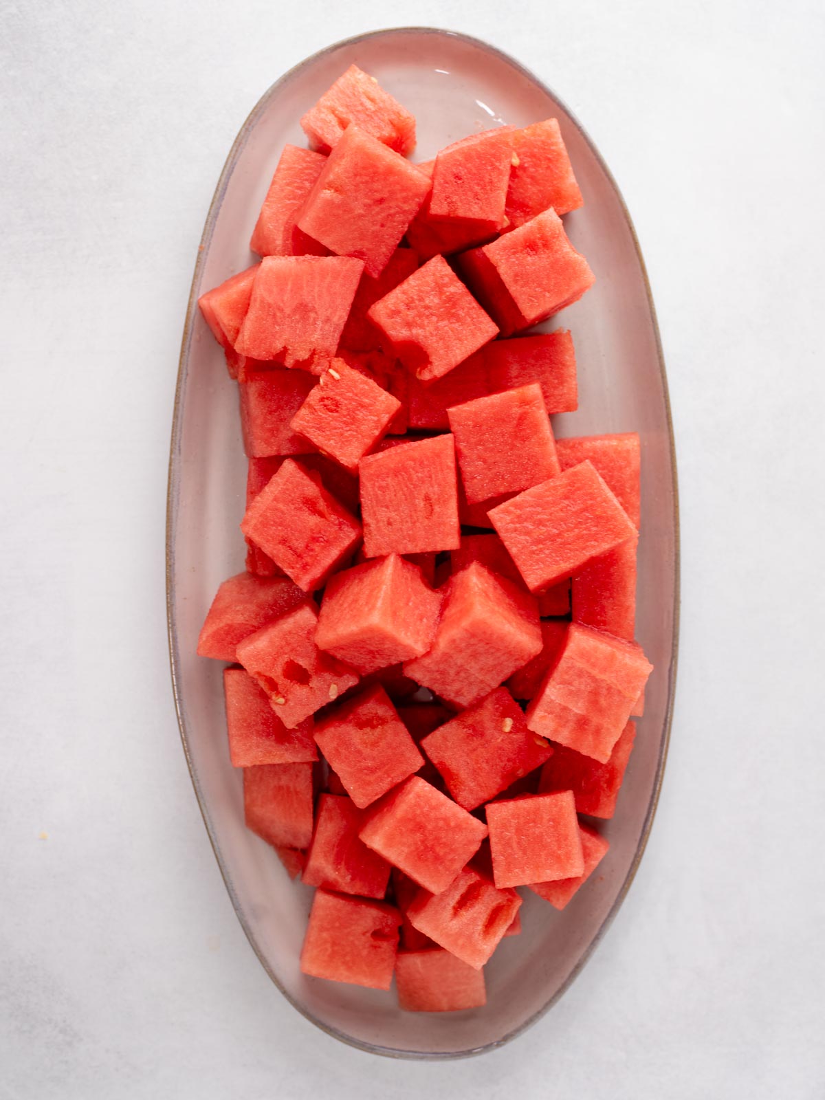 A clear oval plate filled with evenly cut cubes of fresh, red watermelon, placed on a light-colored surface.