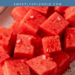 Cubes of fresh watermelon on a plate, sprinkled with Tajin seasoning and sea salt, with lime halves nearby.