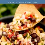 A close-up of a tortilla chip scooping black bean corn dip, with visible corn, black beans, red bell pepper, onion, and herbs—capturing the heart of every great Corn Dip. Text above reads Black Bean Corn Dip and sweetlysplendid.com on a blue banner.