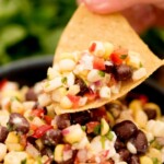 A hand holds a chip with a scoop of flavorful black bean dip, corn, and feta dip above a bowl of the colorful mixture. The text reads black bean corn and feta dip.