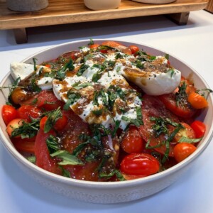 A bowl of caprese salad with sliced tomatoes, cherry tomatoes, fresh mozzarella, chopped basil, and a drizzle of balsamic dressing, set on a white countertop.