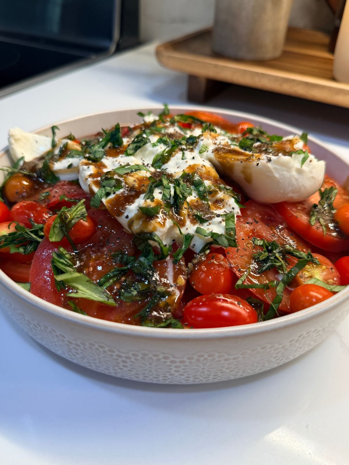 A bowl of tomato salad with mozzarella slices, fresh basil, and a drizzle of balsamic glaze, served on a white countertop.
