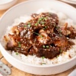 A bowl of white rice topped with braised meat in a dark sauce, garnished with sesame seeds and chopped green onions.