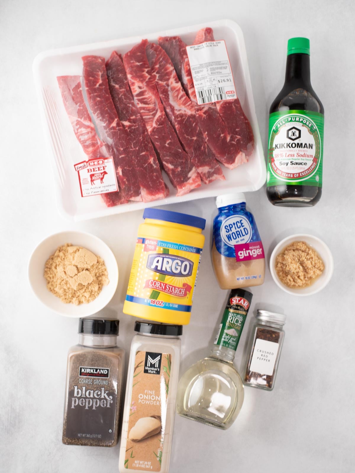Various ingredients are laid out on a white surface, including sliced beef, soy sauce, brown sugar, ground ginger, garlic, cornstarch, black pepper, white wine, and crushed red pepper.