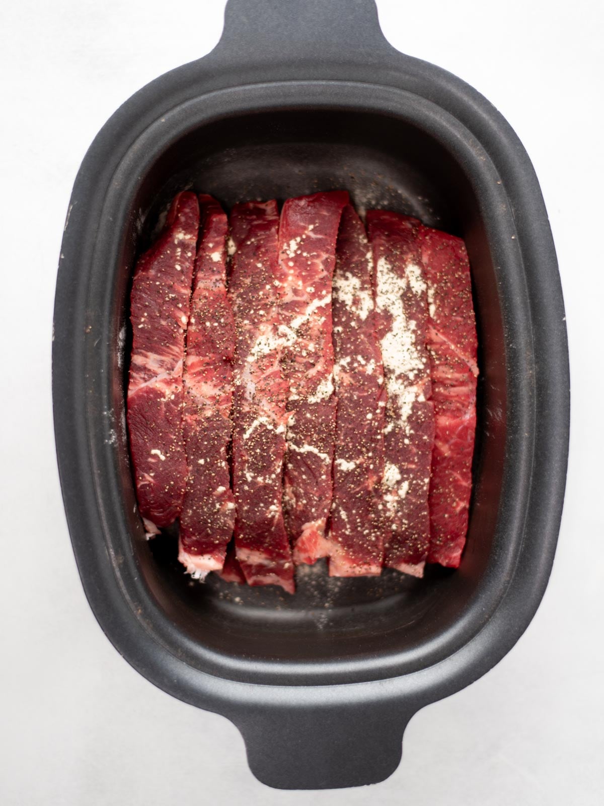 Raw beef slices with seasonings are arranged in a black slow cooker or roasting pan, ready for cooking. The photo is taken from above on a white background.