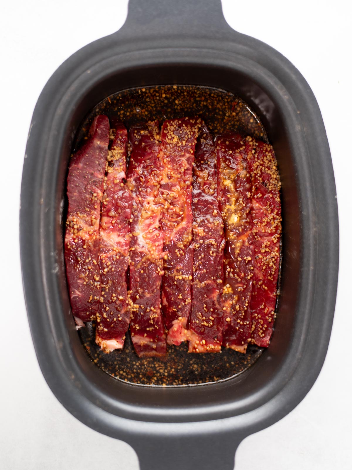Slices of raw beef seasoned with spices are arranged in a black slow cooker, ready to be cooked.