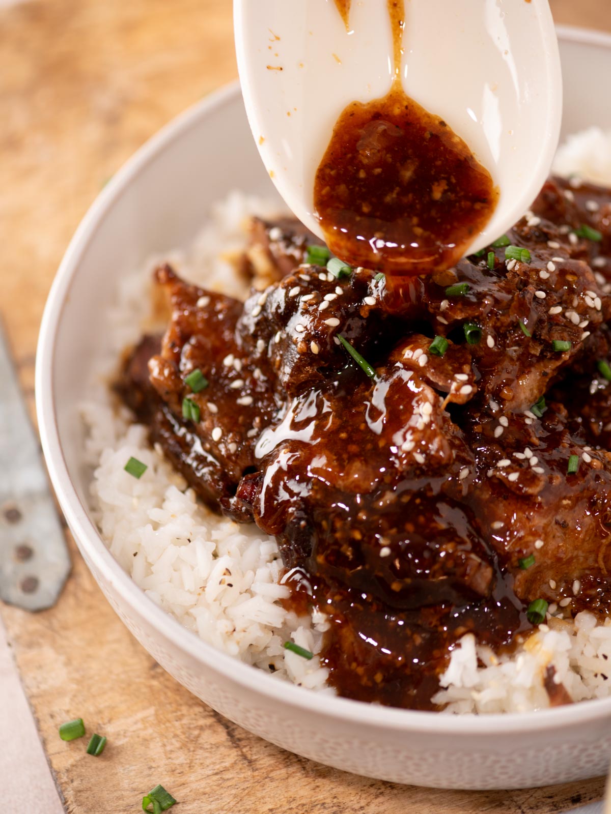 A bowl of white rice topped with glazed beef, sesame seeds, and chopped chives, with extra sauce being poured from a spoon.