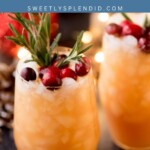 A glass of Christmas punch garnished with cranberries and rosemary sits on a table, surrounded by festive decorations and lights.