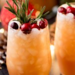 Two glasses of non-alcoholic Christmas punch garnished with cranberries and rosemary sit on a table, with festive decor and lights in the background.