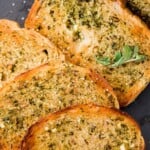 Sliced garlic bread made from sandwich bread arranged on a dark surface, garnished with fresh basil leaves. A loaf of unsliced bread is in the background, and a text overlay reads garlic bread using sliced bread..