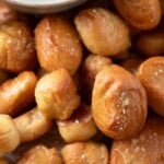 A pile of golden-brown, salted pretzel bites on parchment paper with a bowl of light orange dipping sauce. Text over the image reads soft & chewy pretzel bites and sweetlysplendid.com..