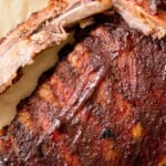 Close-up of smoked ribs with a dark, flavorful crust on parchment paper, garnished with herbs. Text overlay reads award winning smoked ribs and sweetlysplendid.com.