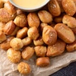 A pile of golden-brown soft pretzel bites sprinkled with salt sits on brown parchment paper, next to a bowl of creamy cheese dipping sauce.