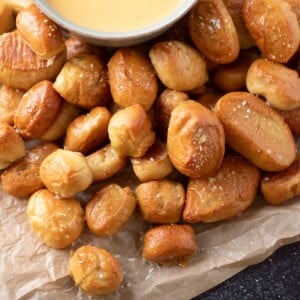 A pile of golden-brown soft pretzel bites sprinkled with salt sits on brown parchment paper, next to a bowl of creamy cheese dipping sauce.