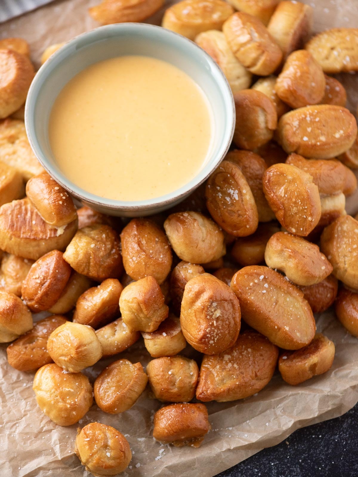 A bowl of cheese dip sits on parchment paper surrounded by salted soft pretzel bites.