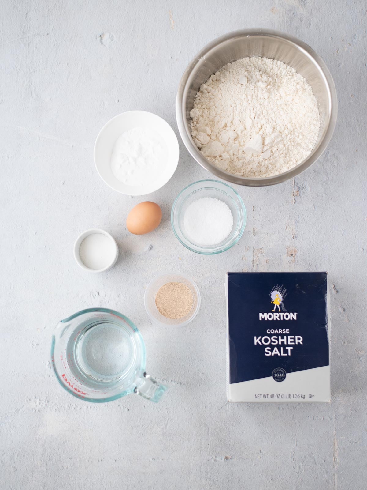 Bowl of flour, bowl of baking powder, egg, bowl of sugar, bowl of yeast, Morton kosher salt box, and glass measuring cup with water on a light surface.