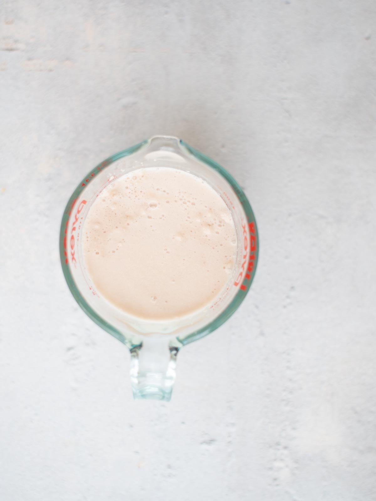 A glass measuring cup filled with a frothy, light beige liquid, viewed from above, placed on a light gray surface.