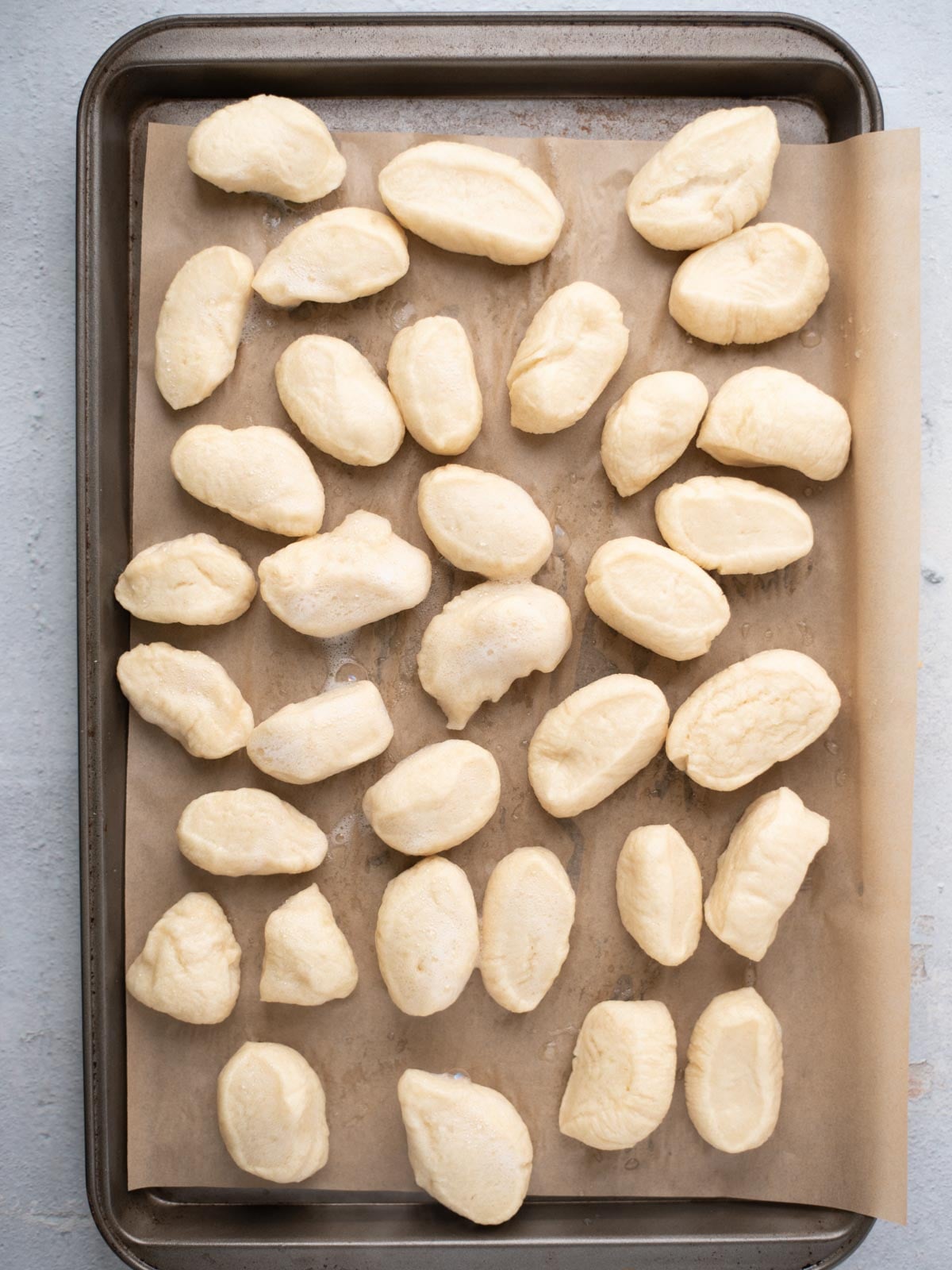 Uncooked gnocchi pieces are arranged on a parchment-lined baking sheet, placed on a light-colored surface, ready to be cooked.