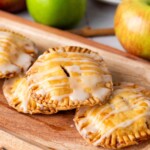 Three mini apple hand pies drizzled with white icing on a wooden board, with fresh apples in the background.