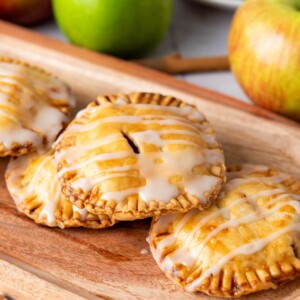 Three mini apple hand pies drizzled with white icing on a wooden board, with fresh apples in the background.