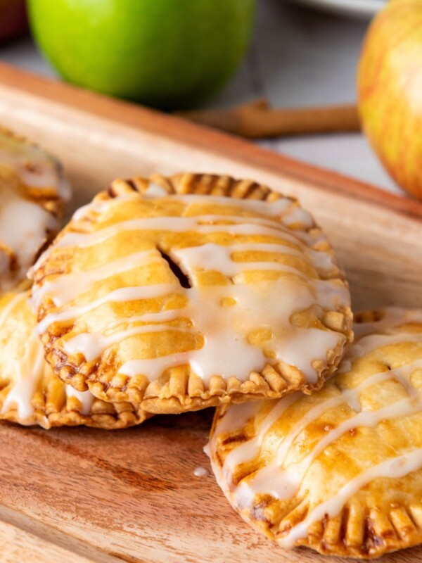 Three mini apple hand pies drizzled with white icing on a wooden board, with fresh apples in the background.