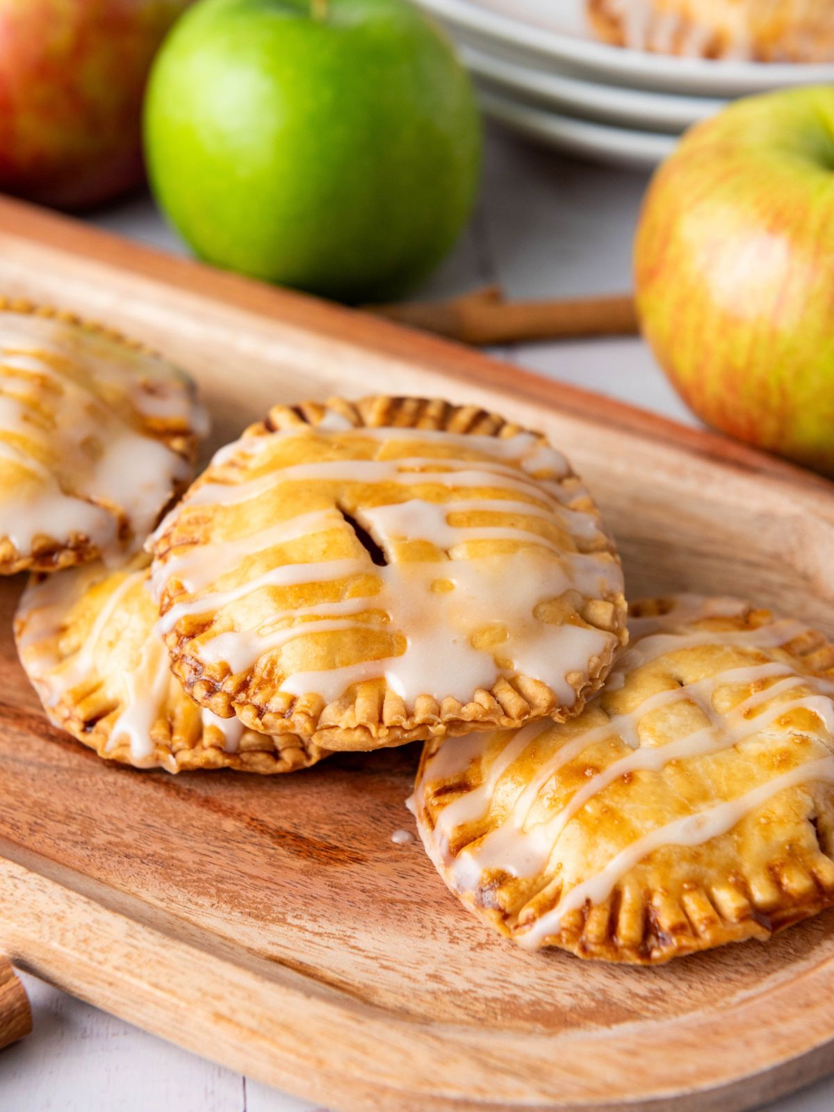 Three small, round hand pies drizzled with white glaze on a wooden board, with two whole apples in the background.